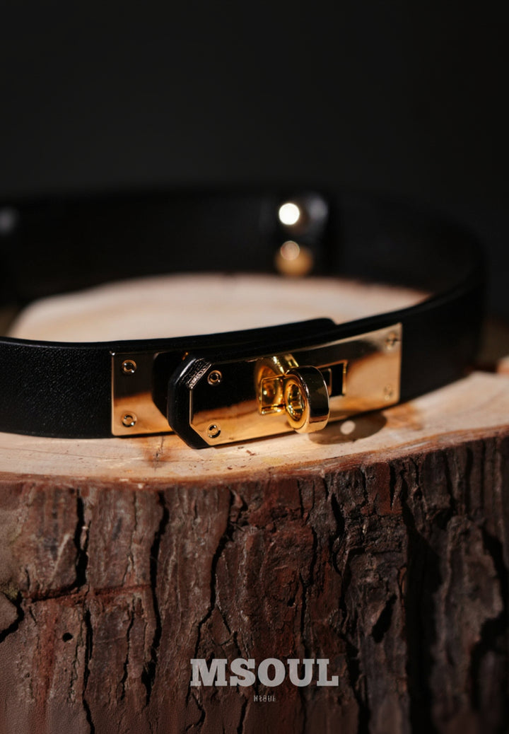 Black leather choker with gold clasp on a wooden block, featuring the brand &